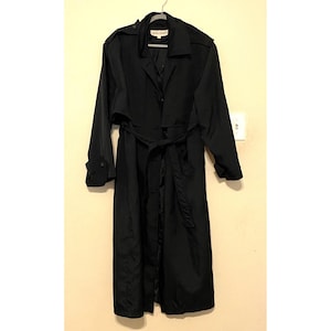 May include: A black trench coat with a belt, hanging on a hanger. The coat has a classic design with a collar, shoulder epaulets, and long sleeves. The coat is made of a smooth, possibly water-resistant material.