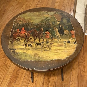 May include: Round wooden side table with a detailed hunting scene painted on the tabletop. The artwork depicts riders on horseback, hounds, and figures in red coats. The table has a dark wood finish and three slender legs.