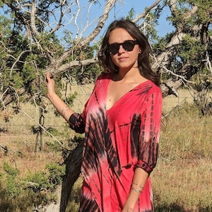 May include: A woman wearing a red and black tie-dye shirt with a deep V-neck, black jeans, and brown cowboy boots. She is standing in a field with a tree behind her.