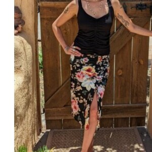 May include: A woman wearing a black tank top and a floral skirt with a slit up the side. She is standing in front of a wooden gate with a sunflower in her hair.