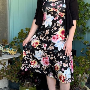 Puede incluir: Una mujer con un vestido negro floral, un c&aacute;rdigan negro y sandalias azul claro. Est&aacute; de pie frente a una pared verde con plantas en macetas.