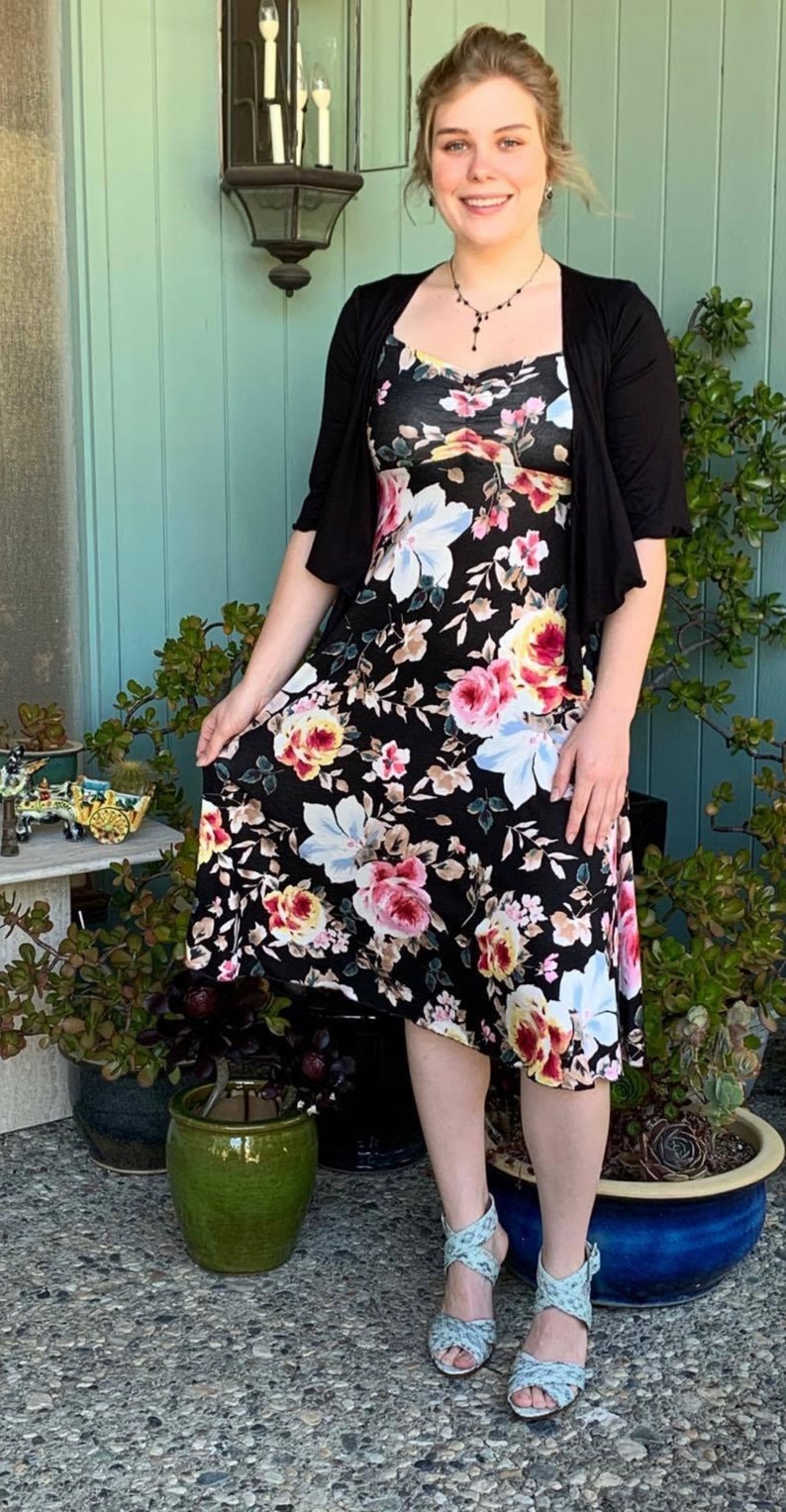 Puede incluir: Una mujer con un vestido negro floral, un c&aacute;rdigan negro y sandalias azul claro. Est&aacute; de pie frente a una pared verde con plantas en macetas.