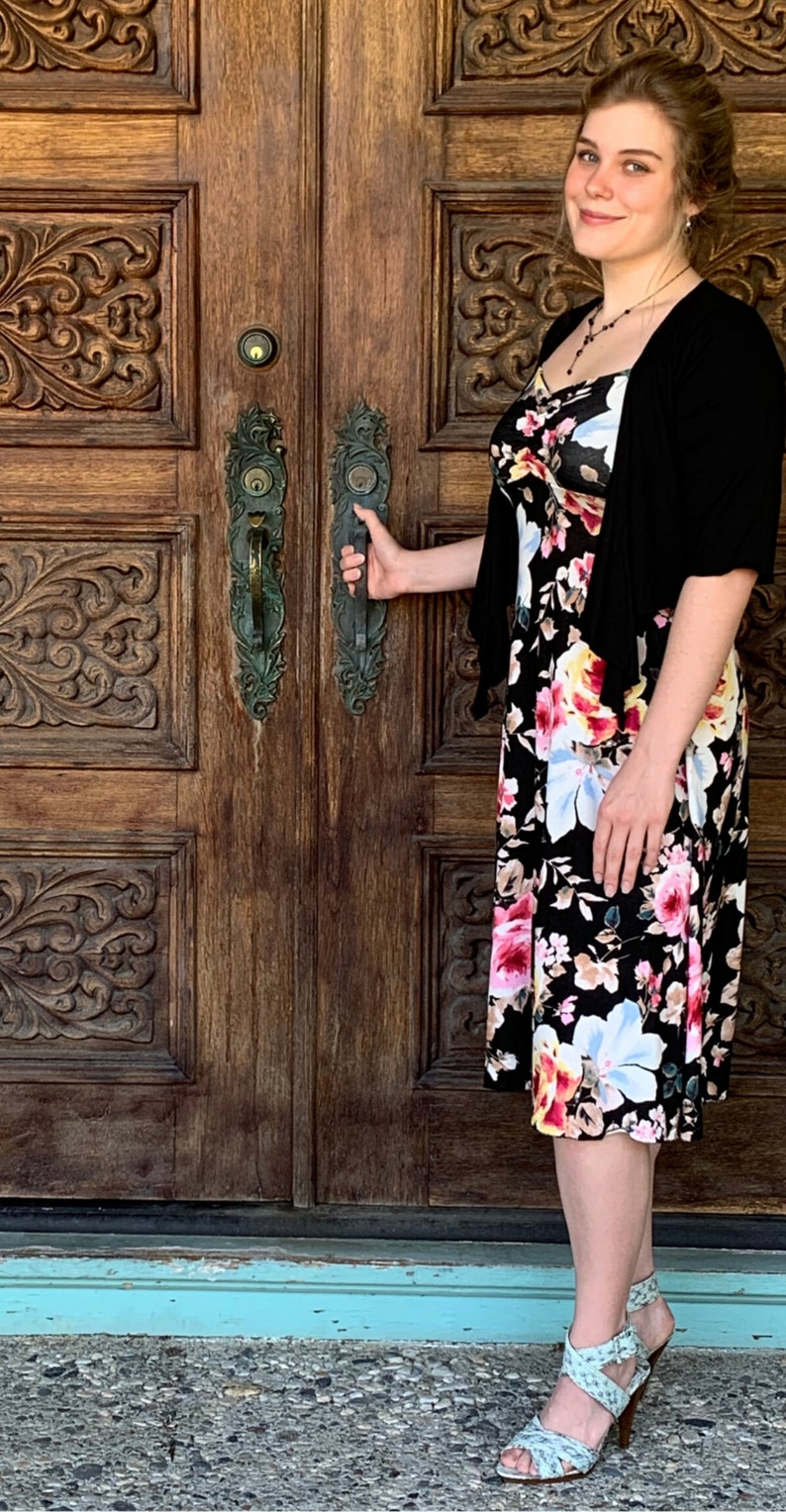 Puede incluir: Una mujer con un c&aacute;rdigan negro y un vestido floral con fondo negro abre una puerta de madera con tallas ornamentadas. La puerta tiene dos manijas de lat&oacute;n y un pomo de lat&oacute;n.