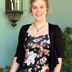 Puede incluir: Una mujer con un vestido negro de flores y un c&aacute;rdigan negro. Est&aacute; de pie frente a una pared verde con una l&aacute;mpara encima de su cabeza.