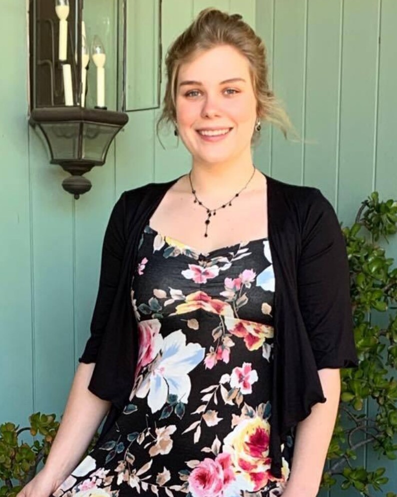 Puede incluir: Una mujer con un vestido negro de flores y un c&aacute;rdigan negro. Est&aacute; de pie frente a una pared verde con una l&aacute;mpara encima de su cabeza.