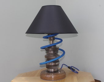 May include: A table lamp with a black lampshade and a base made of stacked metal gears. A blue cord wraps around the gears in a spiral pattern.