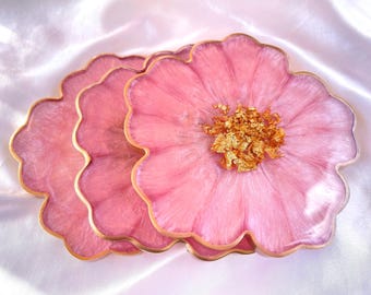 May include: A set of three flower-shaped coasters made of translucent pink resin with a pearlescent finish. Each coaster features a scalloped edge outlined in gold and a textured center filled with metallic gold leaf flakes.
