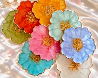 May include: A collection of eight flower-shaped coasters in various colors including red, yellow, green, pink, light blue, dark blue, and white. Each coaster features a scalloped edge and a center filled with gold leaf flakes, creating a textured look.