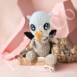 Pode incluir: Um brinquedo de pelúcia de coruja de crochê com pelo branco e marrom, asas pretas e olhos grandes com corações pretos. A coruja está sentada em uma superfície de madeira.