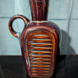 May include: A small, brown ceramic pitcher with a handle and a textured, ribbed design on the front. The pitcher has a glossy finish with hints of blue and orange, and a small opening at the top.
