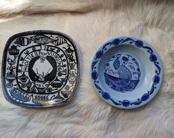 May include: Two vintage ceramic plates with blue and white designs. One plate features a black and white illustration of a chef with the text "Hotel Viking Oslo". The other plate features a blue and white illustration of a person in a boat with fish.