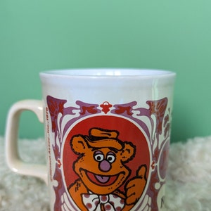 Op de afbeelding: Een witte keramische mok met een bruine beer die een hoed en een strikje draagt. De mok heeft een paars en rood bloemenmotief en de tekst "Copyright Sesame Inc. 1978".