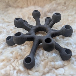 Può includere: Un oggetto decorativo a forma di stella grigio scuro, con più bracci arrotondati, ciascuno terminante con un'apertura circolare. L'oggetto è realizzato in un materiale opaco e poggia su una superficie strutturata bianco sporco. Il design ricorda un fiore stilizzato o una forma geometrica.