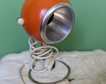 May include: A vintage orange and silver table lamp with a round, orb-shaped shade. The lamp has a white cord and a white switch.