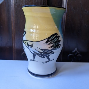 Puede incluir: Un jarrón de cerámica con un esmalte amarillo, azul y blanco. El jarrón presenta un diseño pintado a mano de un pájaro con detalles en blanco y negro.