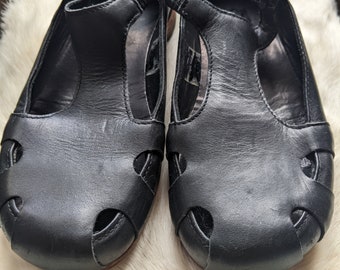 May include: A pair of black leather shoes with a t-strap design and cutouts. The shoes have a rounded toe and a low heel.