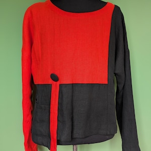 Könnte beinhalten: Ein langärmliges Hemd mit Colorblock-Design. Die obere Hälfte ist leuchtend rot und die untere Hälfte ist schwarz. Ein schwarzer Knopf und ein roter vertikaler Streifen sorgen für Details. Das Hemd wird auf einer schwarzen Schaufensterpuppe präsentiert.