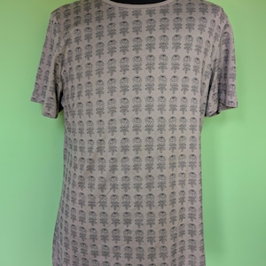 May include: A taupe t-shirt with short sleeves, featuring a repeating pattern of black rose illustrations. The crew neck shirt is displayed on a mannequin. The floral design covers the entire front of the shirt. The background is green.