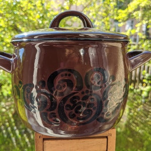 Puede incluir: Una olla de cerámica marrón con tapa y dos asas. La olla tiene un diseño floral negro y marrón.