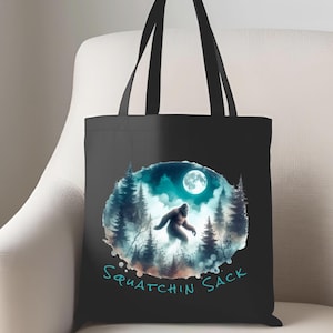 May include: Black tote bag with a Bigfoot illustration set in a forest under a full moon. The words "Squatchin Sack" are written in teal below the image. The bag has black handles.