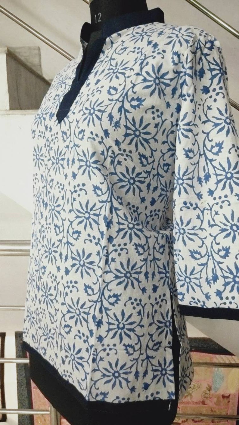 May include: A white cotton tunic with a blue floral print and a black trim. The tunic has a mandarin collar and three-quarter sleeves.
