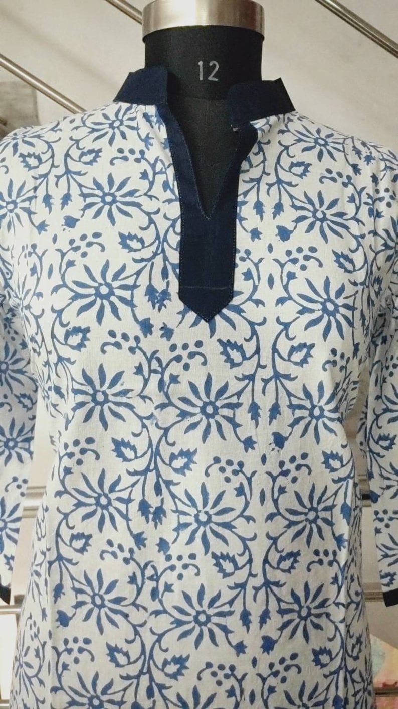 May include: A white cotton tunic with a blue floral print and a navy blue collar. The tunic has a V-neck and three-quarter sleeves.