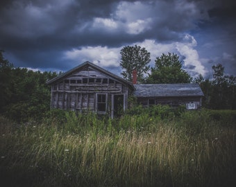 May include: An old, weathered wooden house with a dark, stormy sky overhead. The house is surrounded by overgrown grass and foliage, with a "For Sale" sign visible. The building shows signs of age and disrepair, with missing boards and a dilapidated appearance.