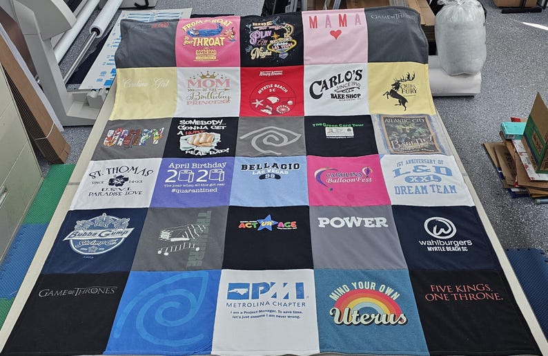 May include: A quilt made from a variety of t-shirts, featuring different designs and text. The quilt is made up of 16 squares, each with a different design. Some of the designs include a heart, a crown, a butterfly, a skull, a rainbow, and a star. The text on the quilt includes the words "Mama", "Mom", "St. Thomas", "April Birthday 2020", "Belllagio", "LSD", "Power", "SPI", "Mind Your Own Uterus", and "Five Kings, One Throne".