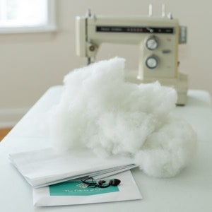 May include: A white sewing machine with a large pile of white stuffing on a white table. The table has a white paper bag and a small card with the text "The Fabric of Life" on it.
