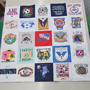 May include: A memory quilt constructed from twenty-five square panels of various soccer-themed t-shirts. Each panel features unique graphic prints, including tournament logos, soccer balls, and text such as AAU Soccer, Tulsa Cup, and Oklahoma Cup.