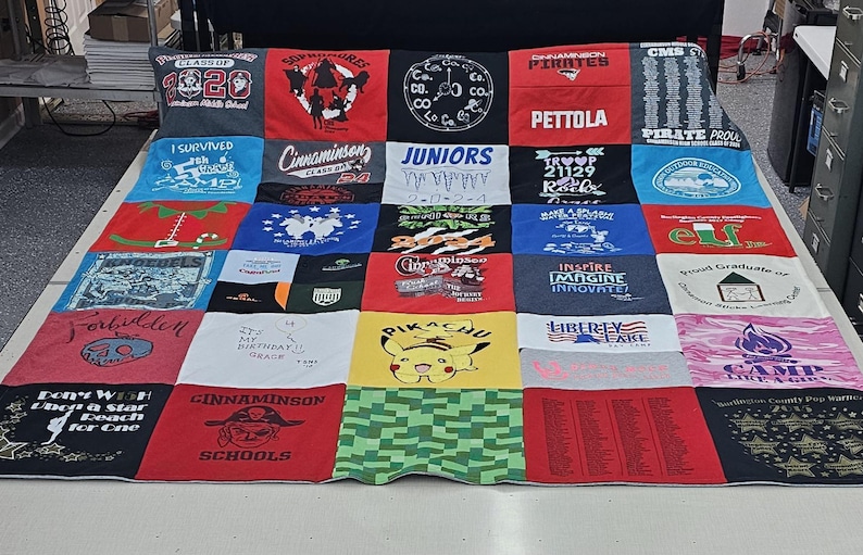 May include: A red, white, and blue patchwork quilt made from t-shirts. The quilt is made up of 25 squares, each with a different design. Some of the designs include the words "Class of 2020", "Cinnaminson", "Juniors", "Pirates", "Pettola", "Forbidden", "My 14th Birthday", "Pikachu", "Inspire Imagine Innovate", "Liberty Lake", "Camp", and "Elf".