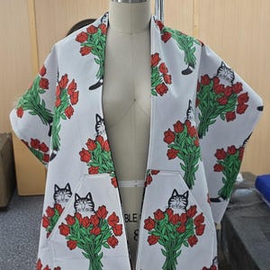 May include: A white open-front jacket featuring a repeating pattern of black and white cats holding bouquets of red tulips with green stems and leaves. The garment includes two front pockets, each displaying a cat graphic among the flowers.