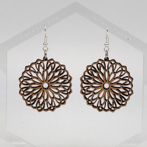 May include: A pair of brown wooden earrings with a delicate, intricate mandala design. The earrings are hanging from silver hooks.