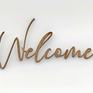 May include: Wooden welcome sign with a cursive font. The sign is brown and has a white background.