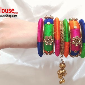 Puede incluir: Un conjunto de pulseras de hilo de seda de colores con detalles dorados y adornos de piedras preciosas. Las pulseras están apiladas en una muñeca y tienen un borla dorada colgante.