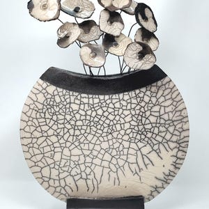 Könnte beinhalten: Dekorative Keramikvase mit runder Form und schwarzem Sockel. Die Vase hat eine rissige, cremefarbene Oberfläche mit schwarzen Linien. Sie hat ein schwarzes Band am oberen Rand und hält eine Gruppe stilisierter, blütenartiger Elemente.