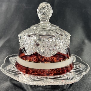 Puede incluir: Una mantequera de cristal transparente con tapa, con una base rojo rubí con una franja blanca y ribete dorado. La tapa y la base están intrincadamente cortadas con patrones de diamantes. El plato se asienta sobre un plato festoneado a juego.
