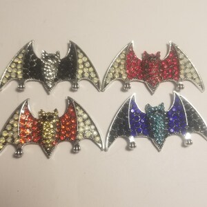 May include: Four silver metal bat charms with different colored rhinestones. The bats have red, blue, black, and yellow rhinestones.
