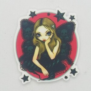 May include: A colorful sticker featuring a fairy with black wings and a red background. The fairy has blonde hair, blue eyes, and is wearing a black dress. The sticker is surrounded by black stars.