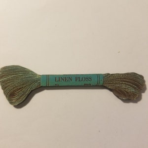 May include: A spool of green linen floss with a blue label that reads "LINEN FLOSS" with the words "No." and "Shade" printed below.