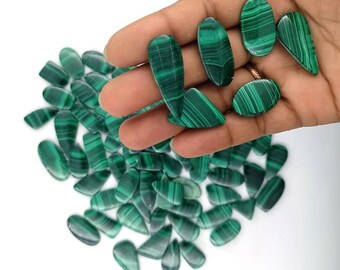 May include: A collection of polished malachite gemstones in various shapes, including ovals and teardrops. The gemstones are a vibrant green with darker green banding. Some stones are held in a hand, while others are scattered on a white surface.