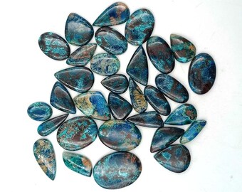 May include: A collection of polished gemstones in various shapes, including oval, teardrop, and irregular forms. The stones display a mix of blue, teal, and brown hues, with a glossy finish. The gemstones are scattered on a white surface.