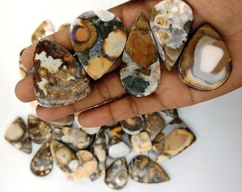 May include: A hand holding a collection of polished gemstones in various shapes. The stones feature a mix of colors, including brown, beige, white, and hints of green. The gemstones are of different sizes and shapes, with a mix of teardrop, oval, and irregular forms.
