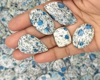May include: Five blue and white speckled gemstone cabochons in various shapes, including teardrop and square, on a hand.