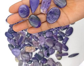 May include: A hand displaying a selection of polished purple charoite gemstones in various shapes, including oval, teardrop, and rectangular. The gemstones show a range of purple hues and textures, with a pile of similar stones in the background.