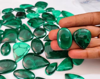 May include: A collection of polished malachite gemstones in various shapes, including ovals and teardrops. The gemstones display a rich, dark green color with swirling patterns. Two stones are held in a hand, showcasing their glossy surface.