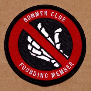 May include: A red and black embroidered patch with a white skeleton hand in a "hang loose" gesture crossed out by a red line. The text "Bummer Club Founding Member" is written in white around the edge of the patch.