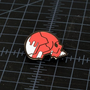 May include: A red and white enamel pin shaped like a skull with a cracked design.