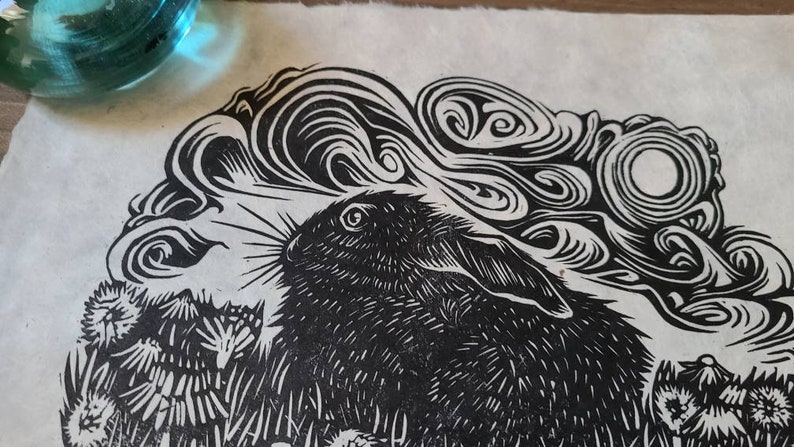May include: Black and white linocut print of a rabbit with swirling clouds and a full moon in the background.
