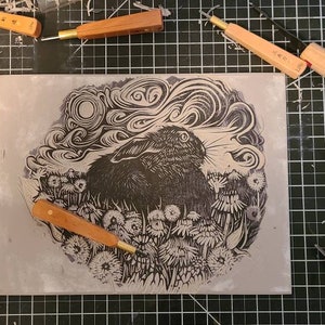 May include: A black and white linocut print of a rabbit in a field of dandelions. The rabbit is looking to the right, and the moon is visible in the sky above. The print is surrounded by carving tools on a black grid.